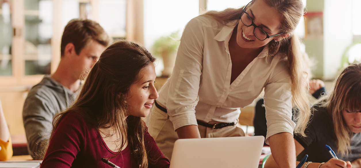 Eine Lehrerin mit Brille und weißem Hemd lächelt, während sie einem Schüler mit langen Haaren am Laptop hilft. Der Schüler schreibt und blickt zur Lehrerin auf. Andere Schüler sind im Hintergrund unscharf zu sehen.