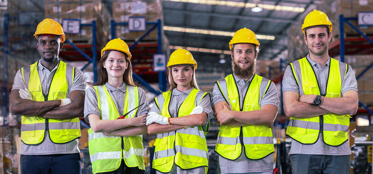 Fünf Lagerarbeiter*innen mit gelben Schutzhelmen und Sicherheitswesten stehen selbstbewusst mit verschränkten Armen da. Sie befinden sich im Innenbereich, im Hintergrund sind Regale mit Kisten vollgestopft.